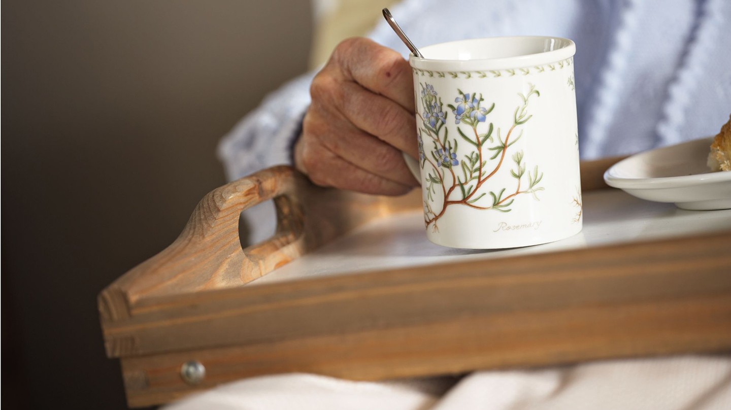 Häusliche Pflege: 7 natürliche Mittel bei leichten Beschwerden Häusliche Pflege mit Heilpflanzen und Hausmitteln: Fotoausschnitt eines Pflegebedürftigen, der im Bett ein Tablett mit einer großen Tasse Kräutertee hält.