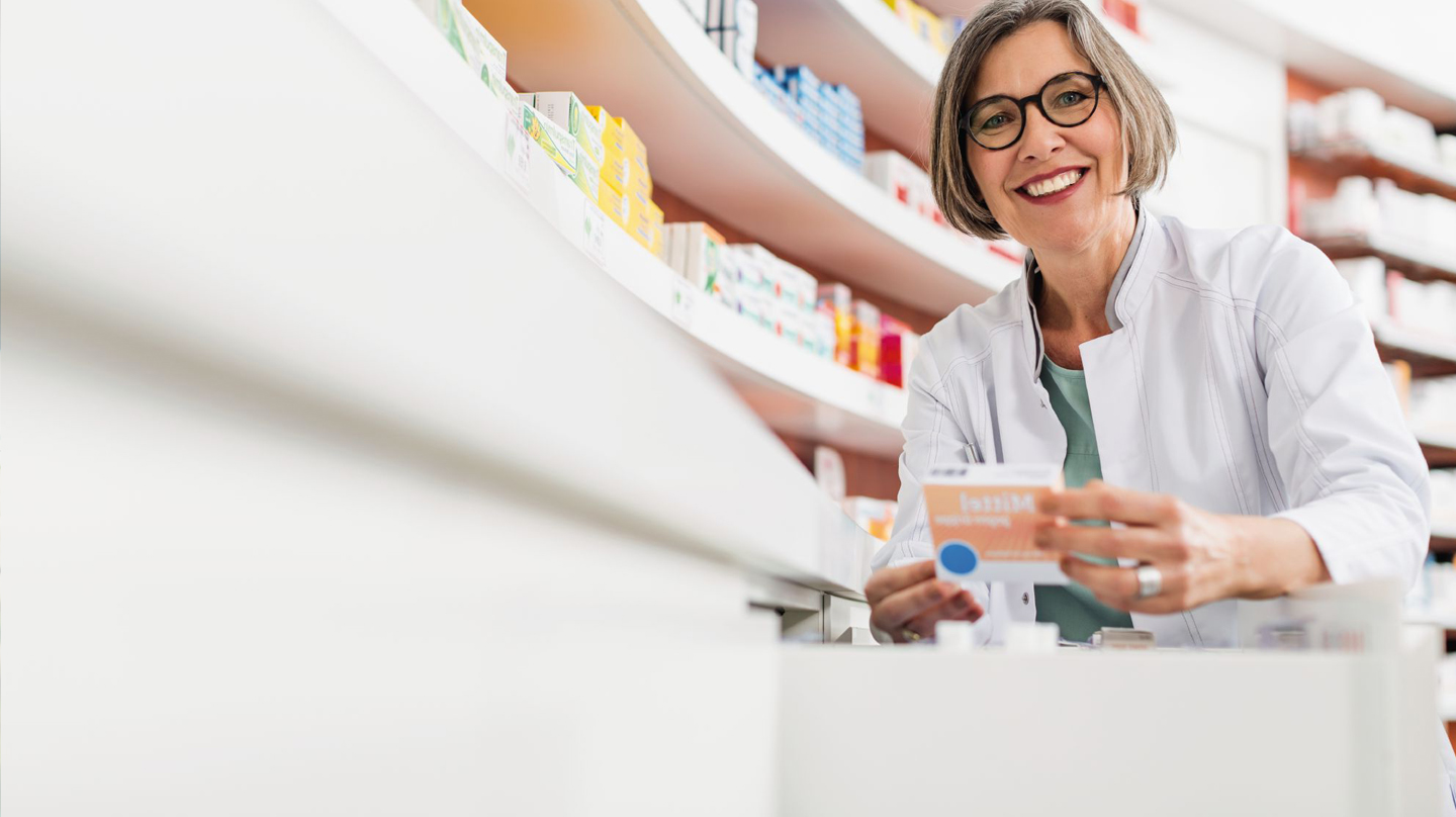 Gefälschte Arzneimittel – wir schützen Sie Arzneimittelsicherheit: Apothekerin vor Regal mit Medikamenten hält eine Medikamentenpackung in der Hand.